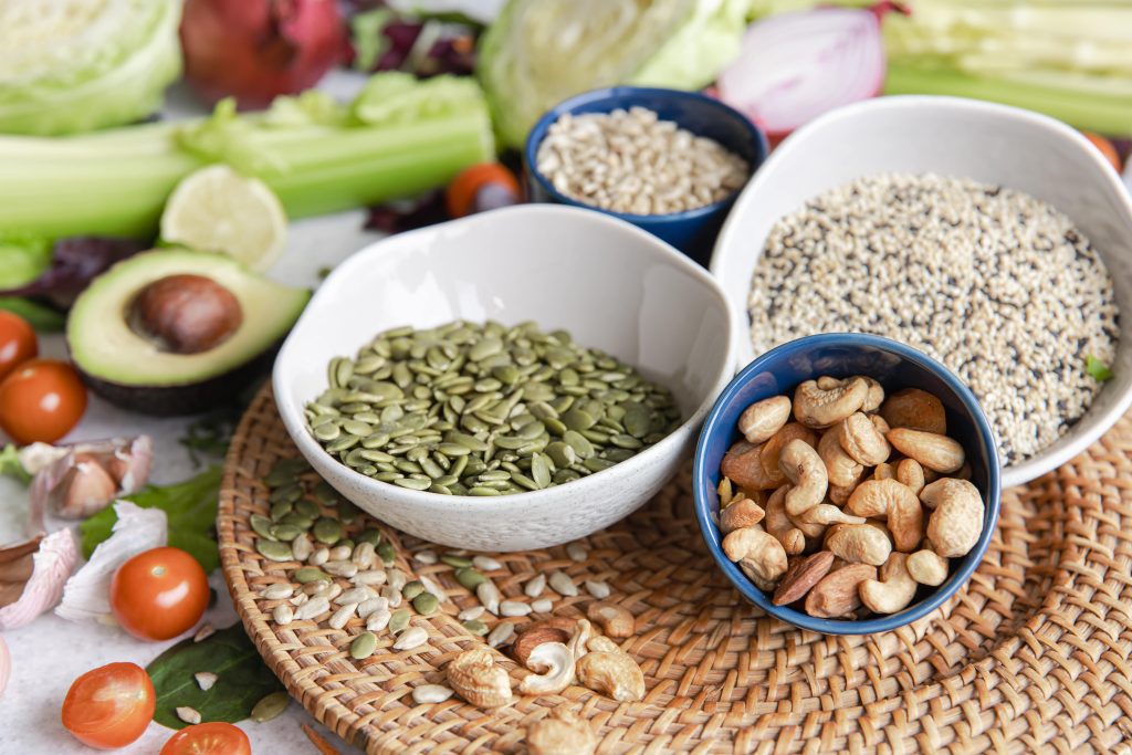 A bowls of pumpkin seeds, cashew nuts, and sesame surrounded by various types of vegetable and plants,